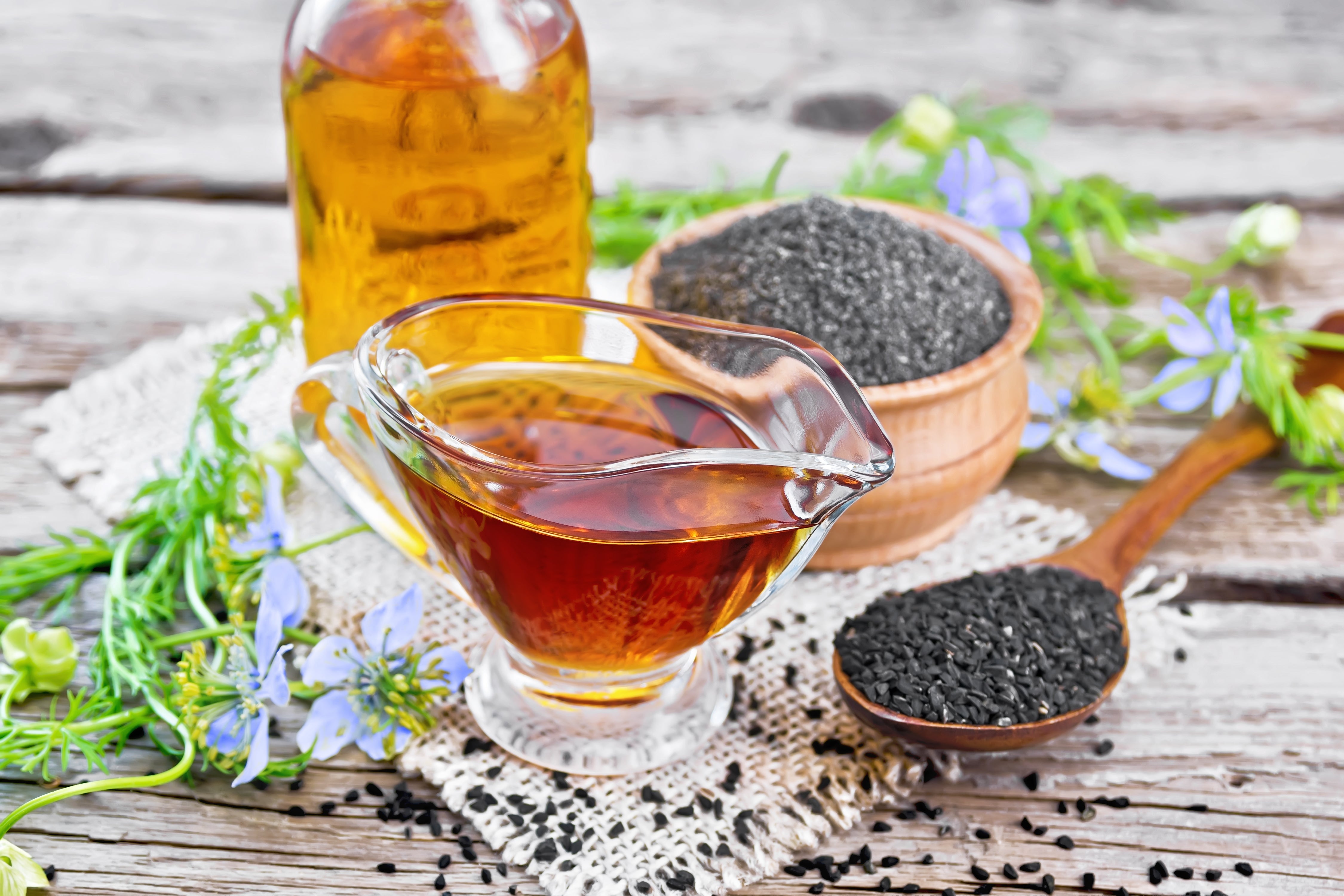 Black seed oil in jar with black seeds and nigella sativa flower on wooden table.